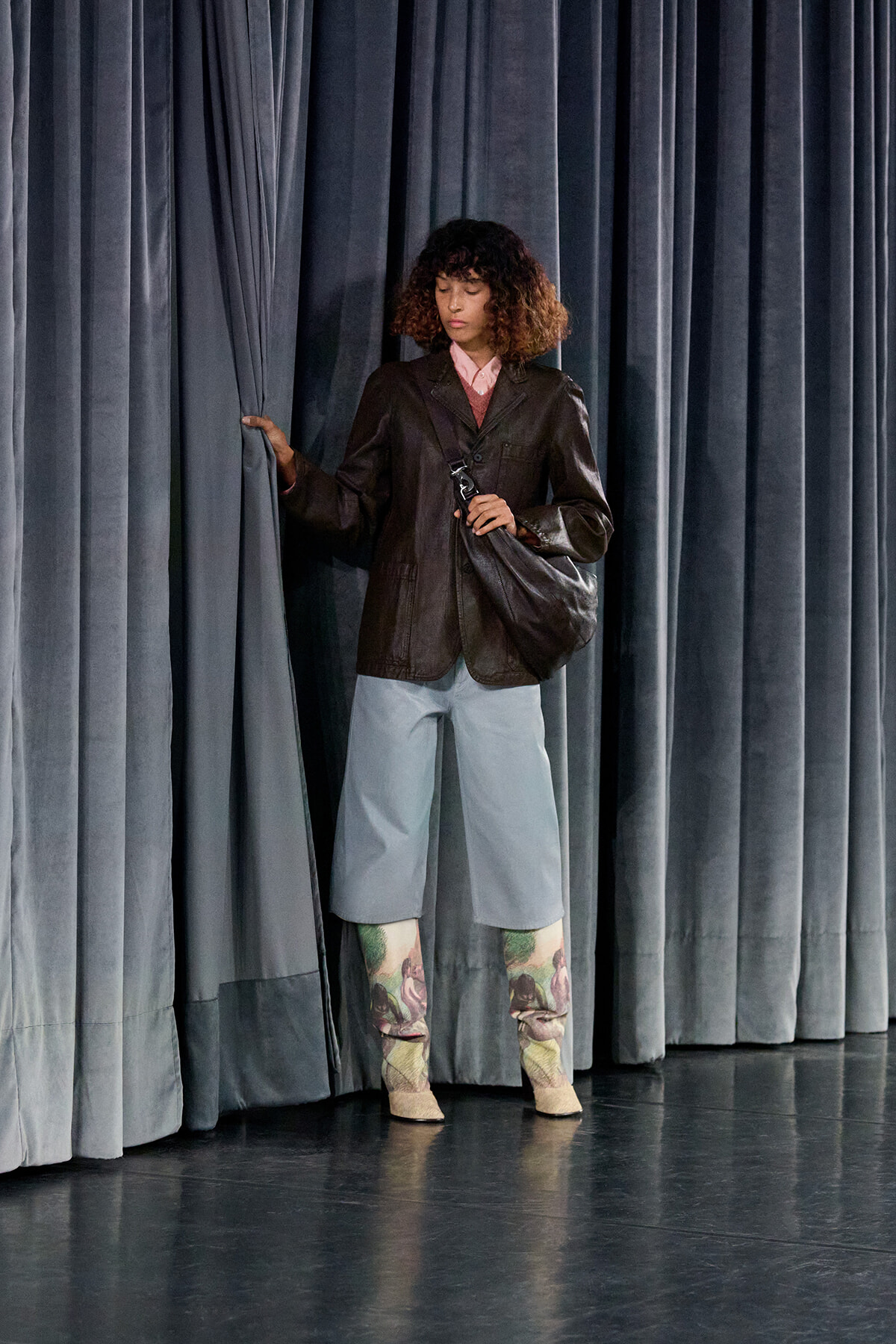 Person standing behind blue stage curtains, wearing a brown leather jacket, pink shirt, light blue wide pants, patterned leggings, and beige boots, holding a bag.