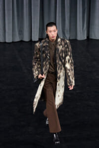Model on a runway wearing a long faux leopard coat over a brown shirt and trousers with black boots and a shoulder bag.