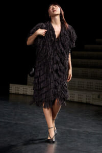 Model walking on stage in a black fringed dress, posing with one hand near the chest and wearing strappy heels.
