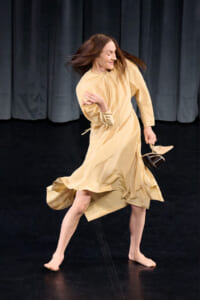 Woman dancing barefoot on a dark stage, wearing a flowing beige dress and holding her shoes in one hand as she spins/moves gracefully.