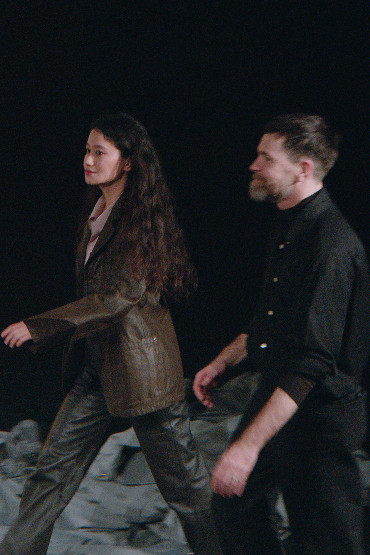 Two adults walk side by side on a dark stage; the woman wears a brown leather suit and the man in a black shirt, both looking ahead.