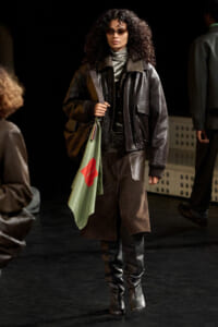 Model walking a runway in a dark leather jacket, turtleneck, and knee-high boots, carrying a light green tote with a red patch and wearing sunglasses.