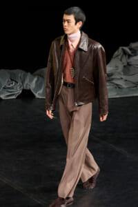 Male model walking on a runway in a brown leather jacket, pink turtleneck, and tan wide-leg trousers with a pendant necklace.
