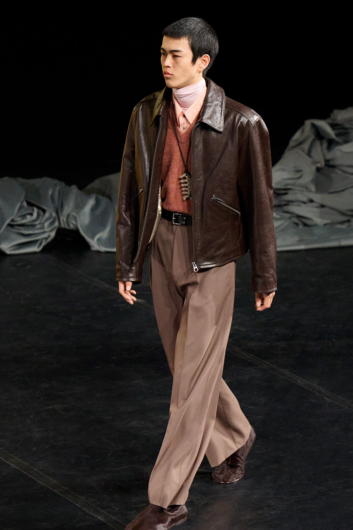 Male model walking on a runway in a brown leather jacket, pink turtleneck, and tan wide-leg trousers with a pendant necklace.