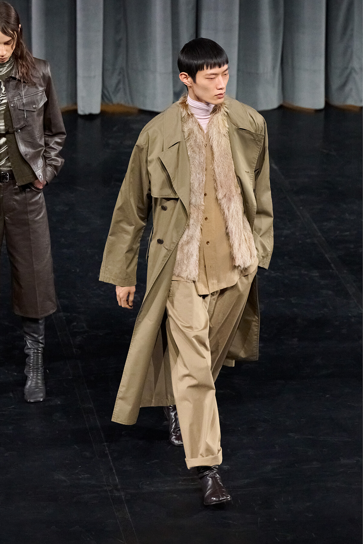 Male model walks a fashion runway in a tan trench coat with fur scarf, tan suit, and brown boots on a dark stage.