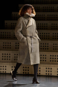 Model walks the runway in a knee-length grey shearling coat with a belt and hands in pockets.