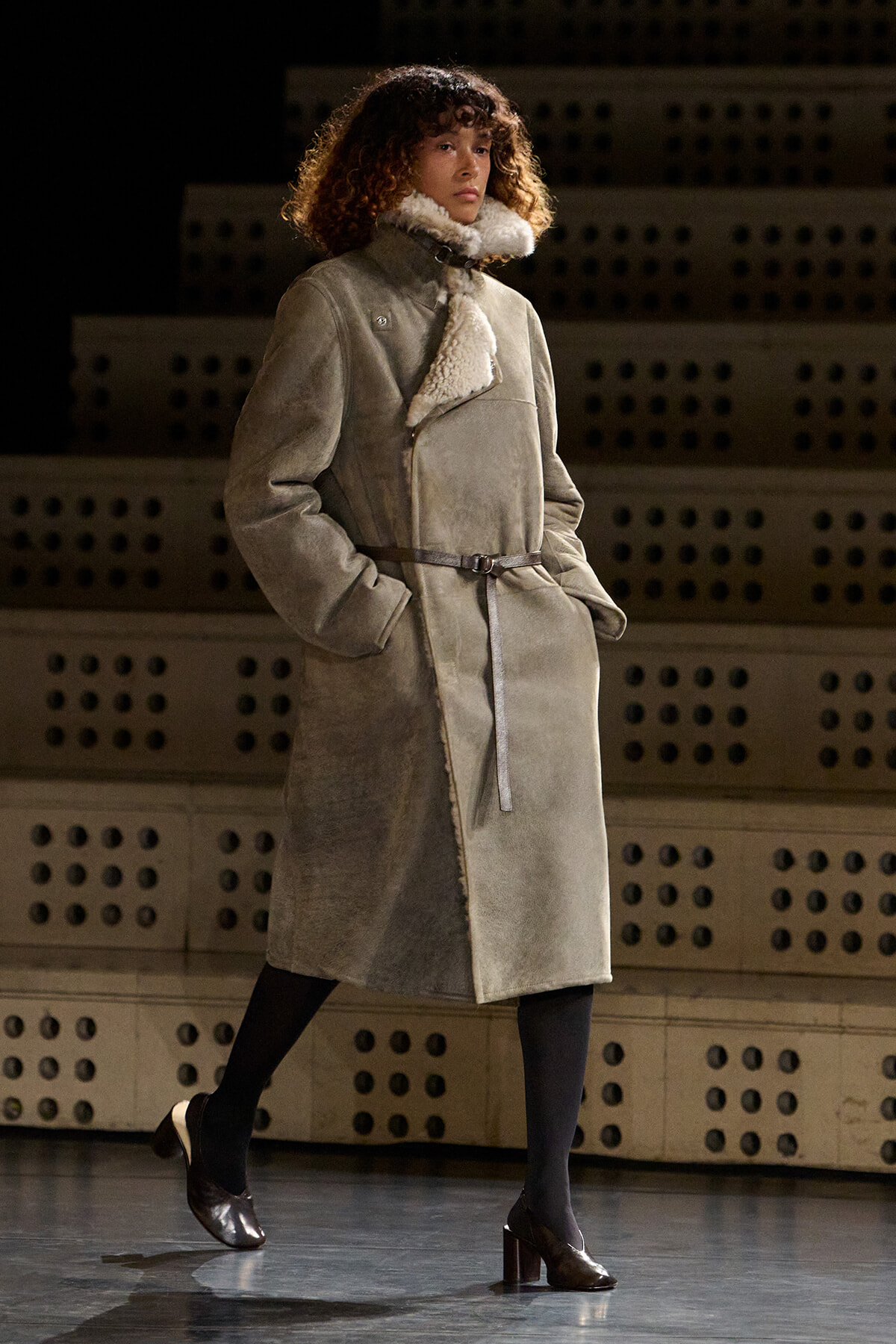 Model walks the runway in a knee-length grey shearling coat with a belt and hands in pockets.