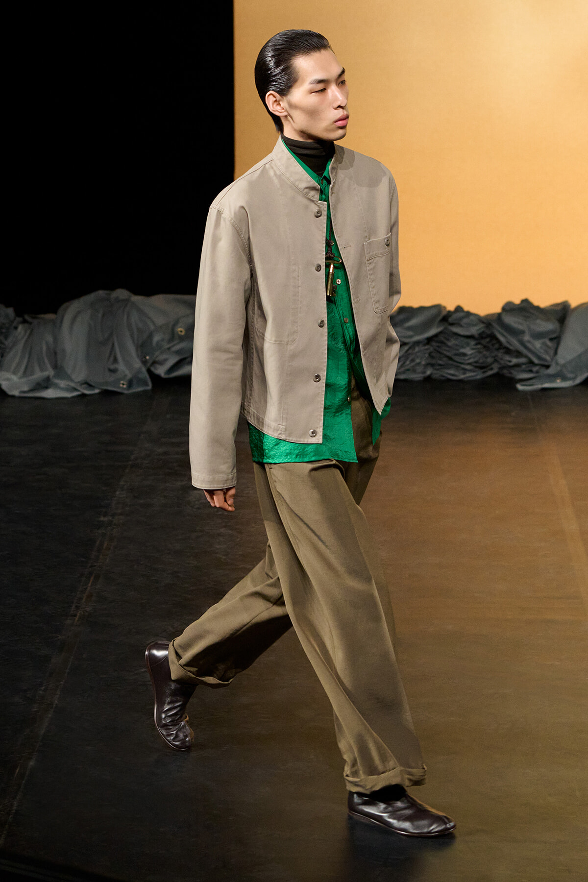 Male model walks a fashion runway in a beige button-front jacket over a green shirt, paired with khaki wide-leg pants and dark brown shoes.