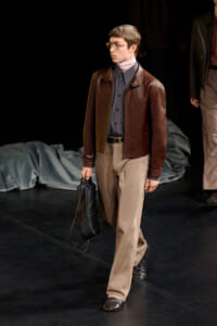Male model in a brown leather jacket, gray shirt, and tan trousers walks the runway while holding a black bag.