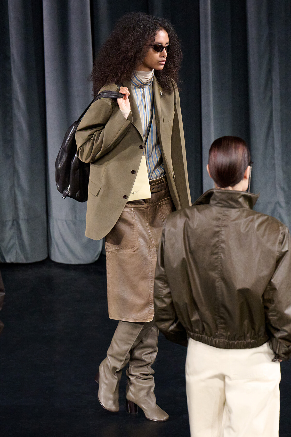 A fashion model with curly hair in a tan blazer, striped turtleneck, brown skirt, and knee-high boots carries a black backpack on a backstage runway setup.