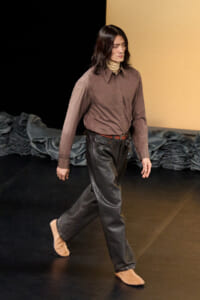 Person with shoulder-length dark hair walks across a stage, wearing a brown button-down shirt, tan turtleneck, black leather pants, and tan slip-on shoes.