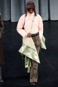 Runway model in a pale pink turtleneck and brown pants wearing a shiny metallic green wrap draped over the shoulder with a crossbody strap and fur hat.