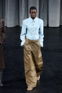 Model walking a runway in a light blue denim shirt and oversized tan-brown wide-leg pants on a dark stage.