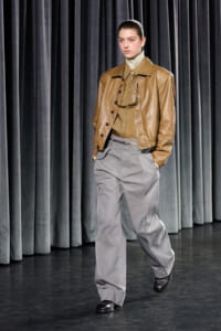 Model walking a runway in a tan leather jacket, beige top, and gray high-waisted pants against dark curtains.