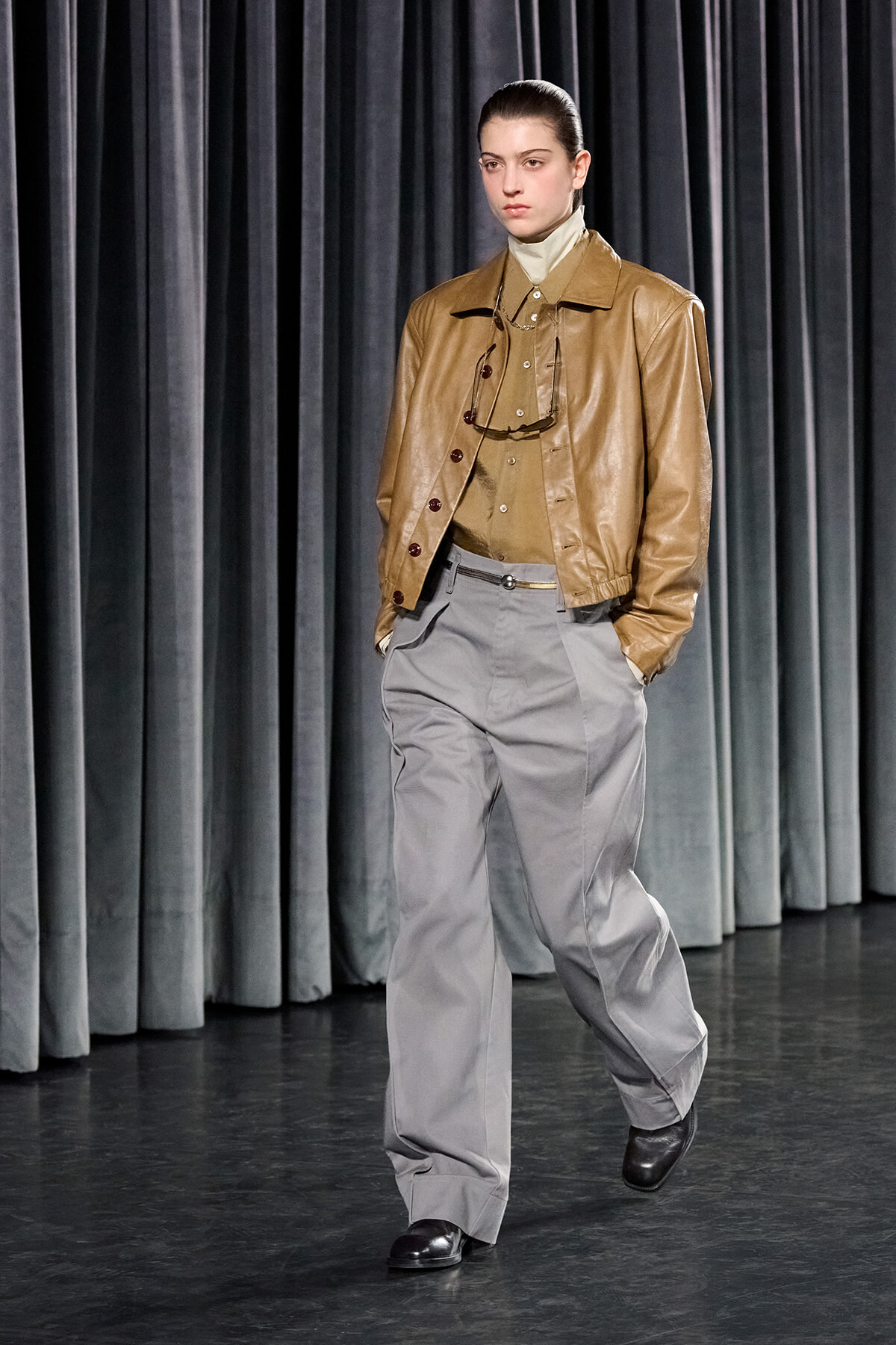 Model walking a runway in a tan leather jacket, beige top, and gray high-waisted pants against dark curtains.