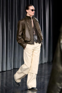 Model walking a fashion runway in a shiny olive-brown bomber jacket over a beige top, paired with wide-leg cream pants and black shoes, wearing slim sunglasses and slicked-back hair.