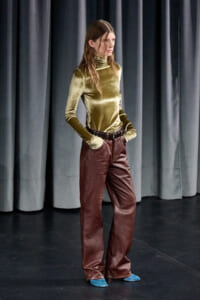 Woman on a stage posing with hands in pockets, wearing a gold velvet turtleneck and brown wide-leg pants with blue shoes behind dark curtains.