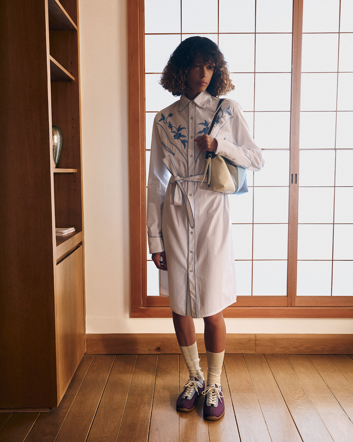 Woman wearing a belted light shirt dress with blue floral embroidery, carrying a pale blue and beige handbag, standing by a wooden-framed shoji screen window in a warm, wood-floored room.