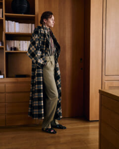Person standing in a wood-paneled room wearing a long blue plaid coat, beige high-waisted trousers, and floral slippers, with hands in pockets.