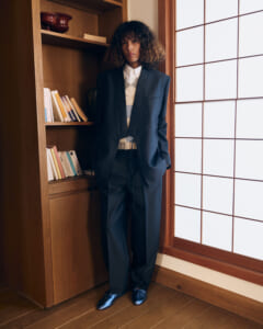 Person in a navy suit standing in a wooden bookshelf alcove beside a frosted grid window, hands in pockets.
