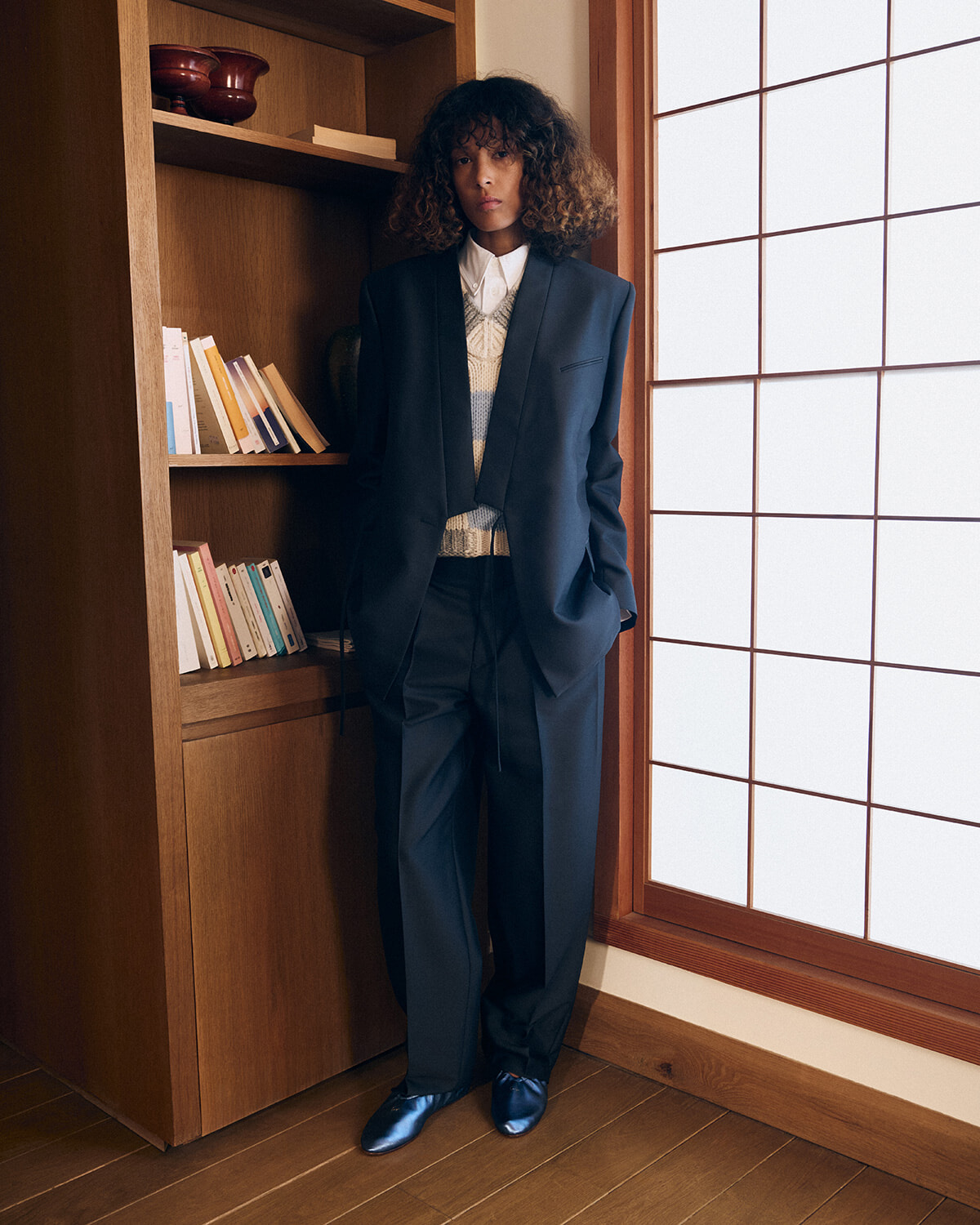 Person in a navy suit standing in a wooden bookshelf alcove beside a frosted grid window, hands in pockets.