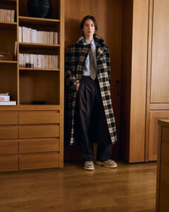 Person stands in a wood-paneled room wearing a long beige-black plaid overcoat, white shirt, tie, wide black trousers, and beige sneakers by a built-in cabinet.
