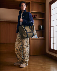 Person in a navy jacket and floral pants stands indoors, carrying a large olive duffel bag.