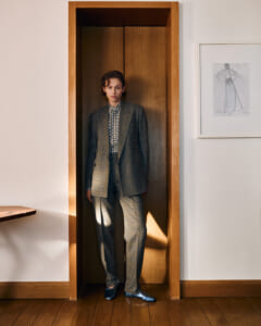 Person in a gray plaid suit and checkered shirt stands in a tall wooden doorway, facing the camera.