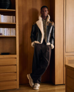 Young man leans against a wooden cabinet wearing a shearling-lined black leather jacket, plaid shirt, dark wide-leg pants, and cream shoes in a home interior.