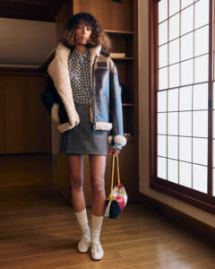 Model wearing a shearling-lined blue jacket over a checkered shirt and gray plaid mini skirt, white socks and cream shoes, carrying a color‑block handbag by a large window.