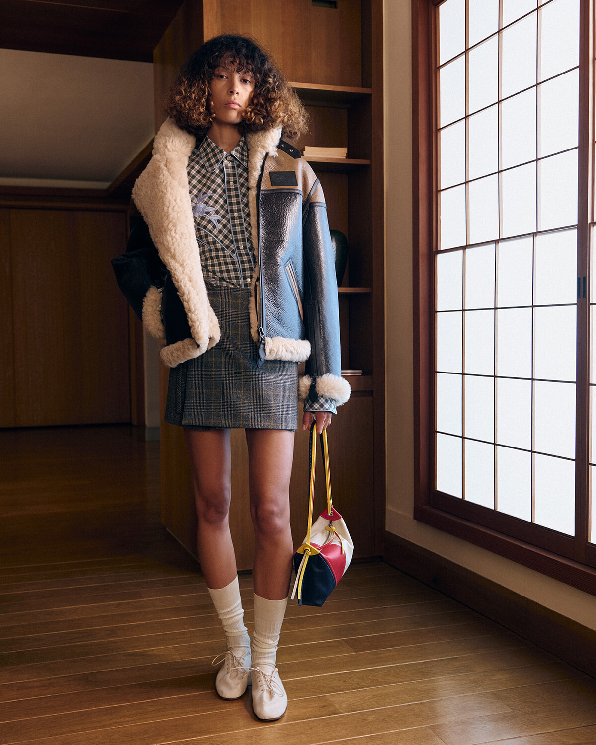 Model wearing a shearling-lined blue jacket over a checkered shirt and gray plaid mini skirt, white socks and cream shoes, carrying a color‑block handbag by a large window.