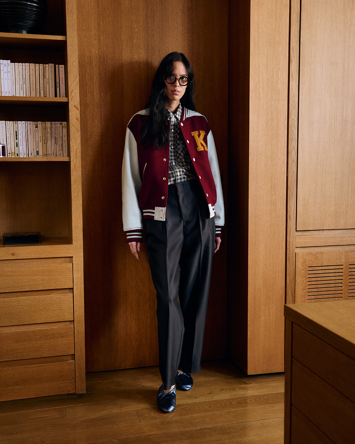 Woman in a burgundy varsity jacket with white sleeves and a yellow 'K' patch, plaid shirt, black wide-leg pants, and blue loafers standing in a wood-paneled room.