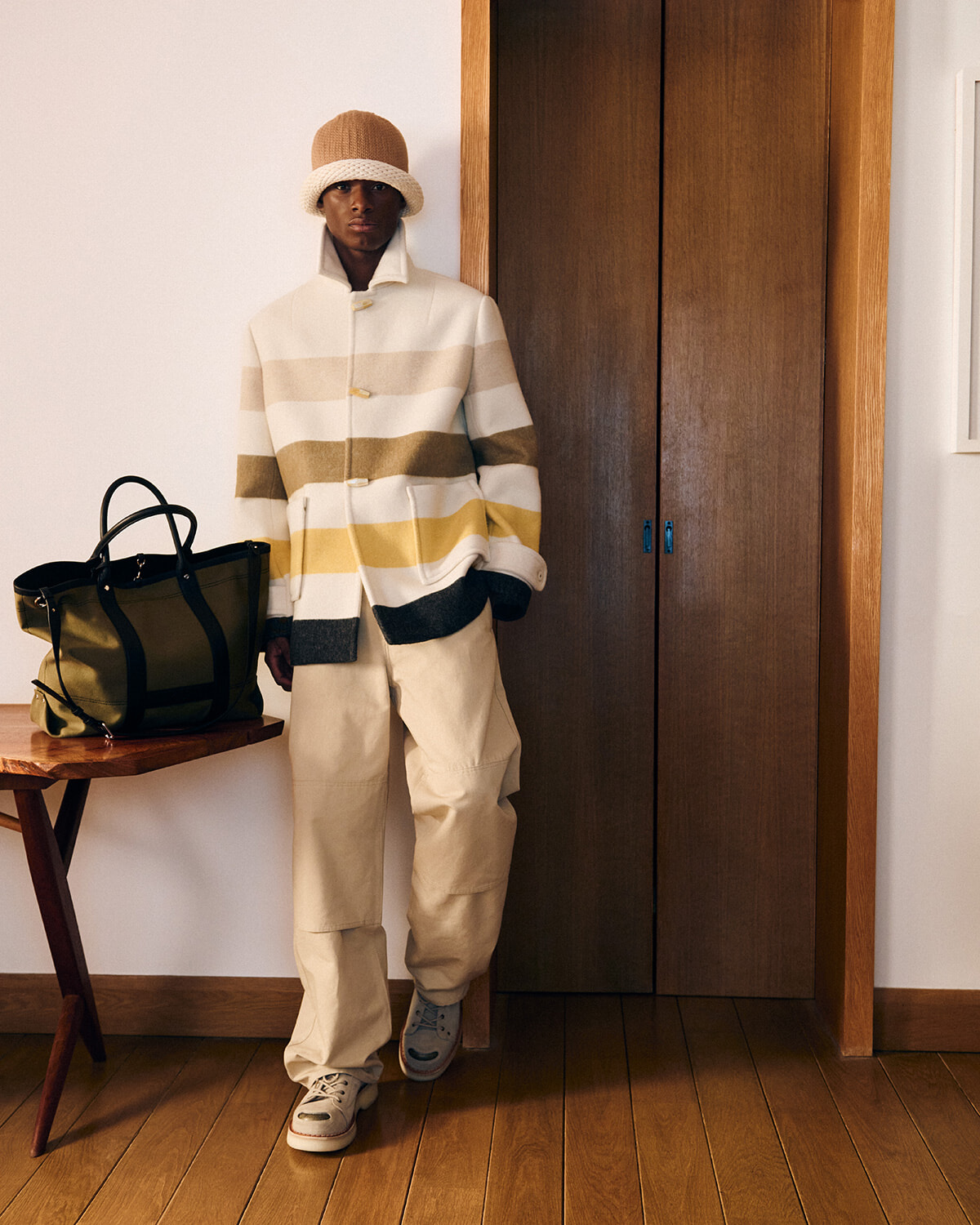 Person wearing a cream and tan striped coat, beige pants, and a tan bucket hat, standing beside a wooden closet with a green bag on a table next to them.