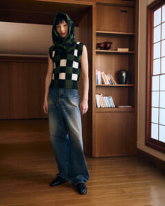 Person wearing a green and white checkered knit hooded vest with loose jeans, standing in a wood-paneled room by a bookshelf.