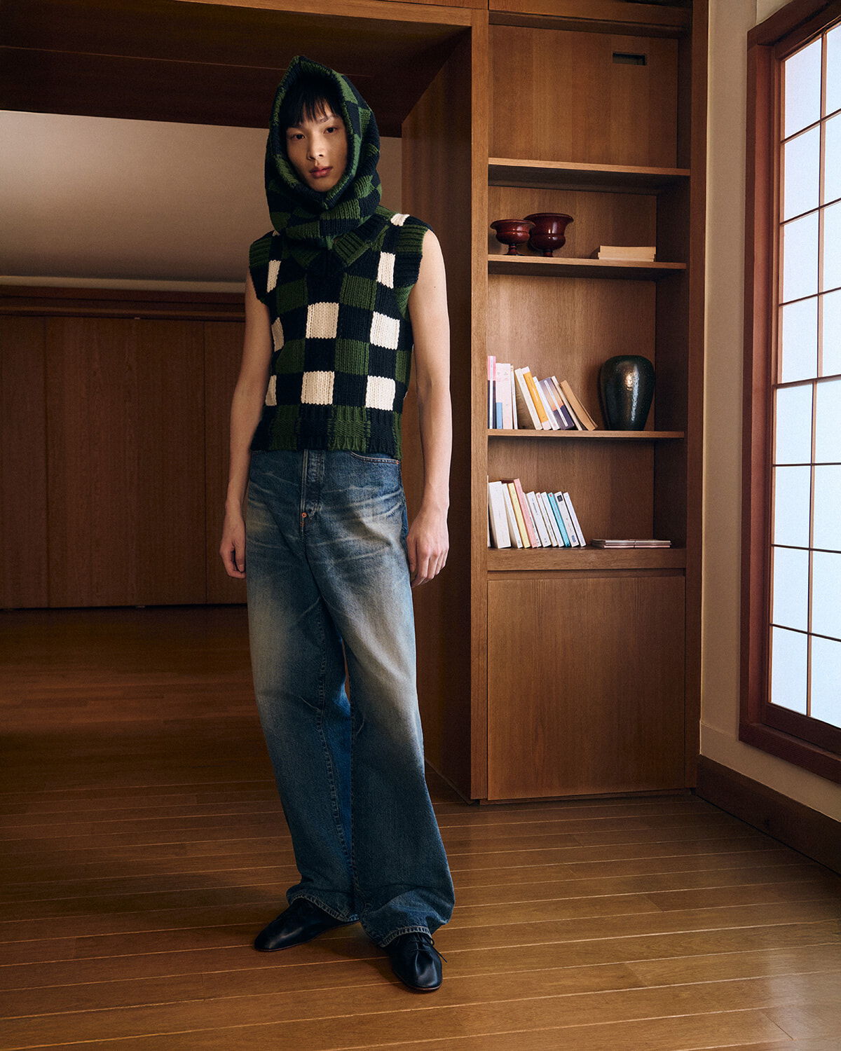 Person wearing a green and white checkered knit hooded vest with loose jeans, standing in a wood-paneled room by a bookshelf.