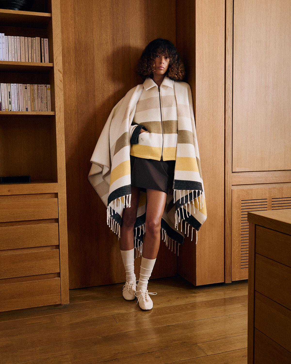 Model wearing a striped poncho with fringe, standing in a warm wood-paneled room with built-in shelves nearby.