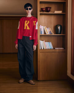 Person wearing a red varsity jacket with yellow 'K' letters, standing beside a wooden bookshelf.