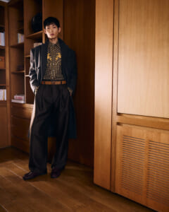 Man wearing a long dark coat and wide trousers, leaning against a wooden panel in a warm, wood-filled room, posing for fashion.