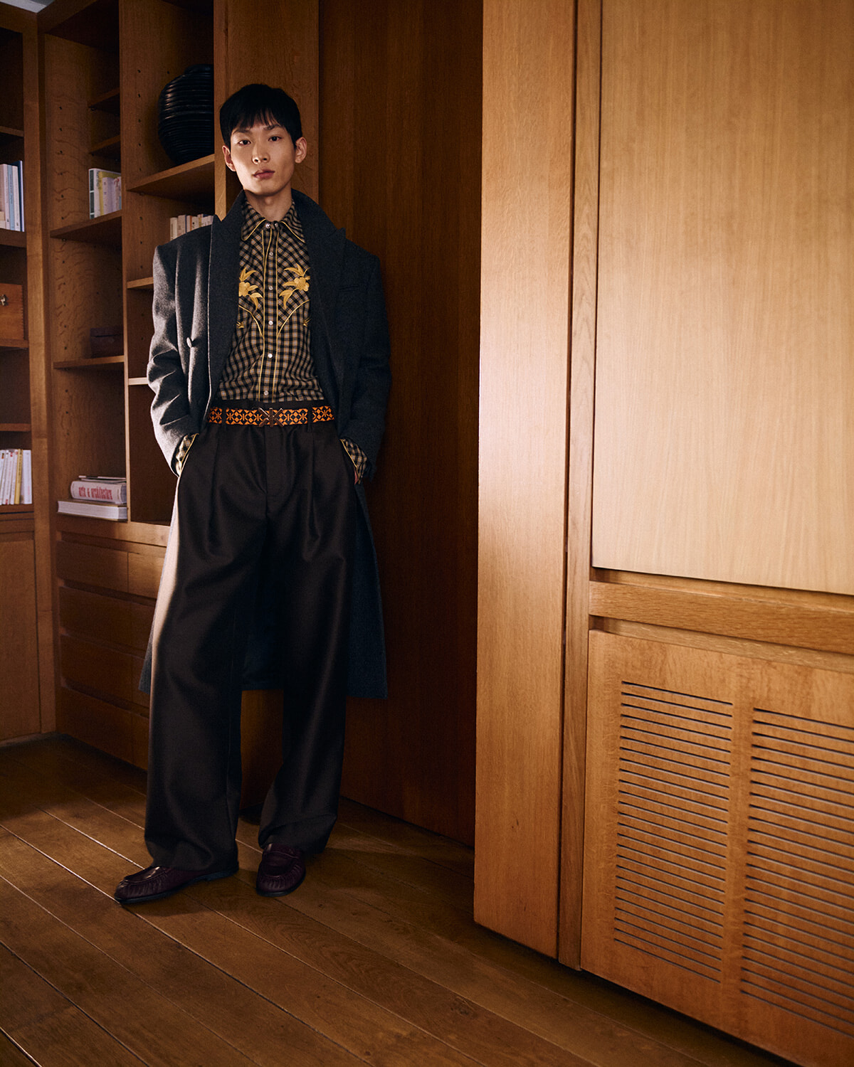 Man wearing a long dark coat and wide trousers, leaning against a wooden panel in a warm, wood-filled room, posing for fashion.