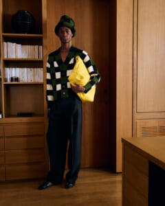 Person wearing a green and black checkered knit cardigan and matching hat, holding a bright yellow bag in a wood-paneled room.