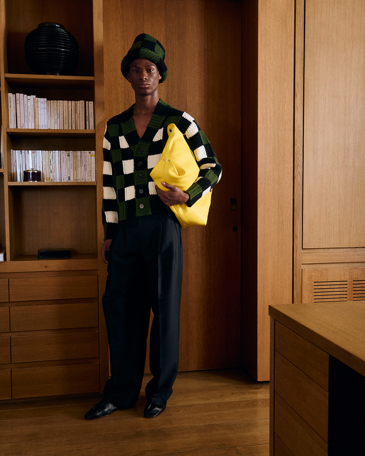 Person wearing a green and black checkered knit cardigan and matching hat, holding a bright yellow bag in a wood-paneled room.