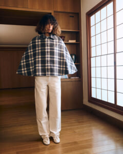 Model wearing a blue plaid cape over cream pants and white shoes in a warm wood-paneled room by a large window.