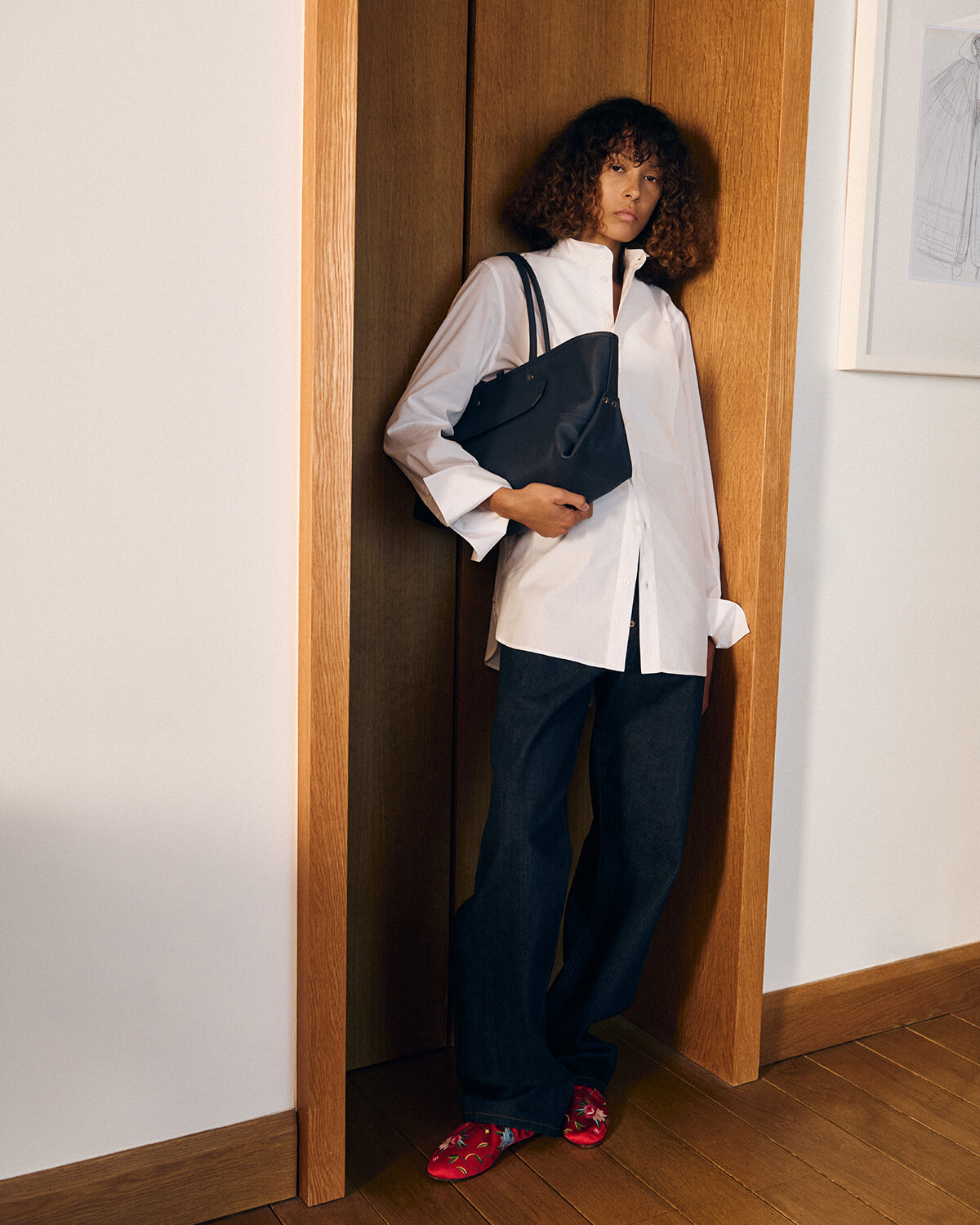 Person leaning in a wood doorway wearing an oversized white shirt, dark wide-leg jeans, red floral slippers, and a black tote bag.