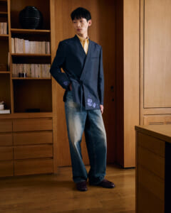 Man posing indoors in a navy embroidered wrap blazer over a yellow shirt, paired with jeans and loafers in a wood-toned room.