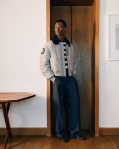 Man poses in a light bomber jacket with striped shirt, hands in pockets in a wooden doorway.