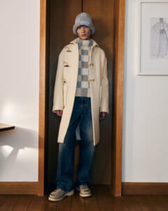 Person wearing a cream duffle coat, gray checkered sweater, jeans, and a knitted hat standing in a doorway.