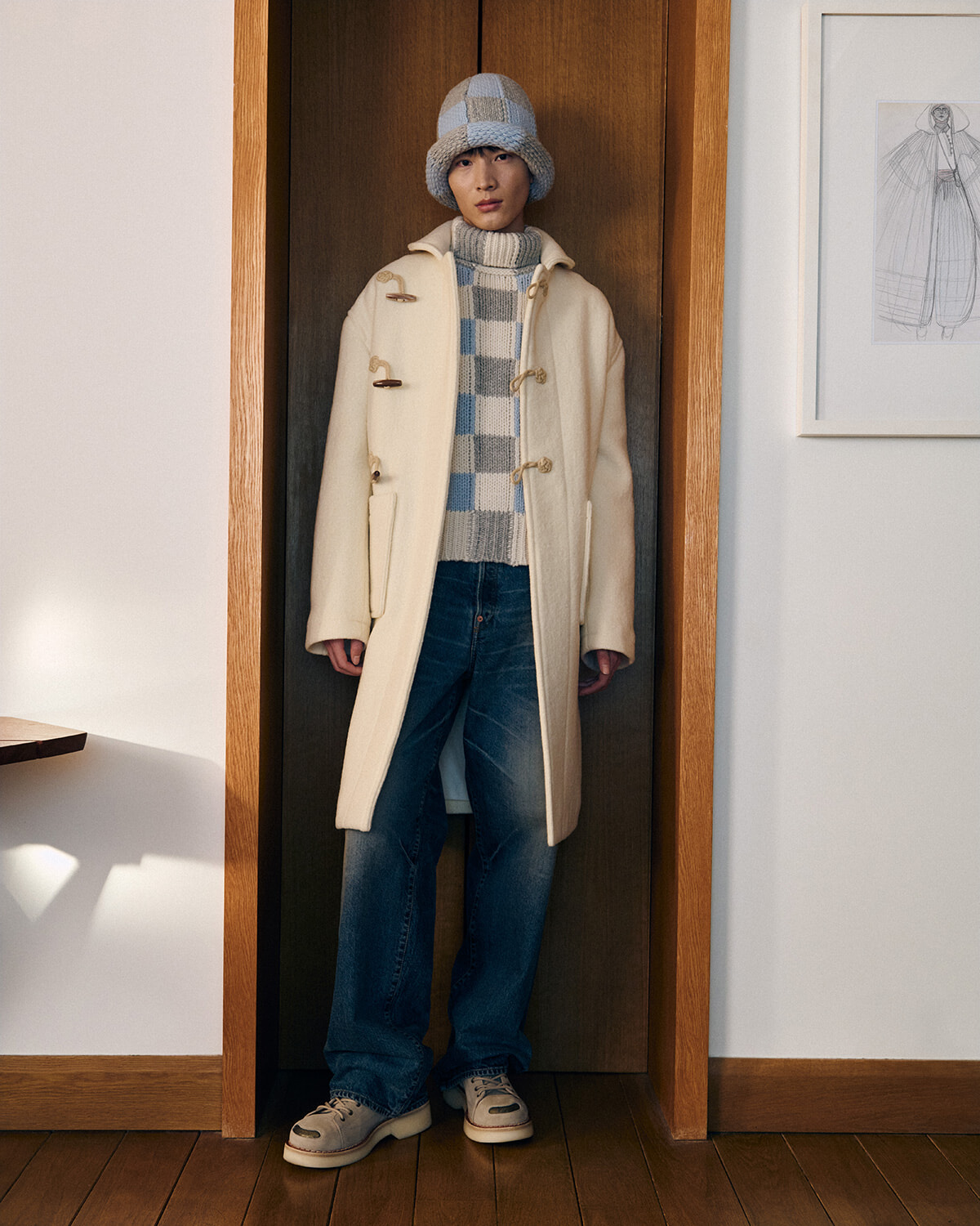 Person wearing a cream duffle coat, gray checkered sweater, jeans, and a knitted hat standing in a doorway.