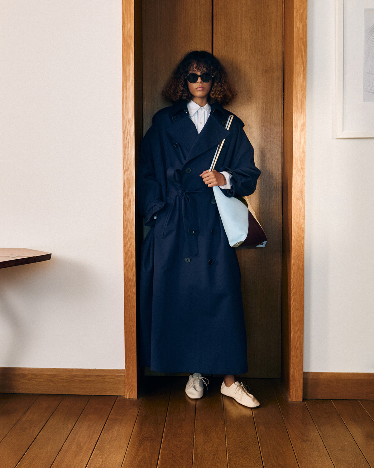 Person wearing a navy trench coat and sunglasses stands in a doorway, holding a light-blue tote bag with a white shirt underneath and white shoes.
