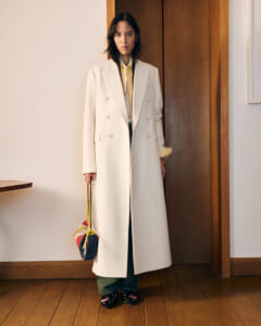 Person standing in a doorway wearing a long cream double-breasted coat, yellow shirt, green pants, and floral shoes, holding a colorful handbag.”,