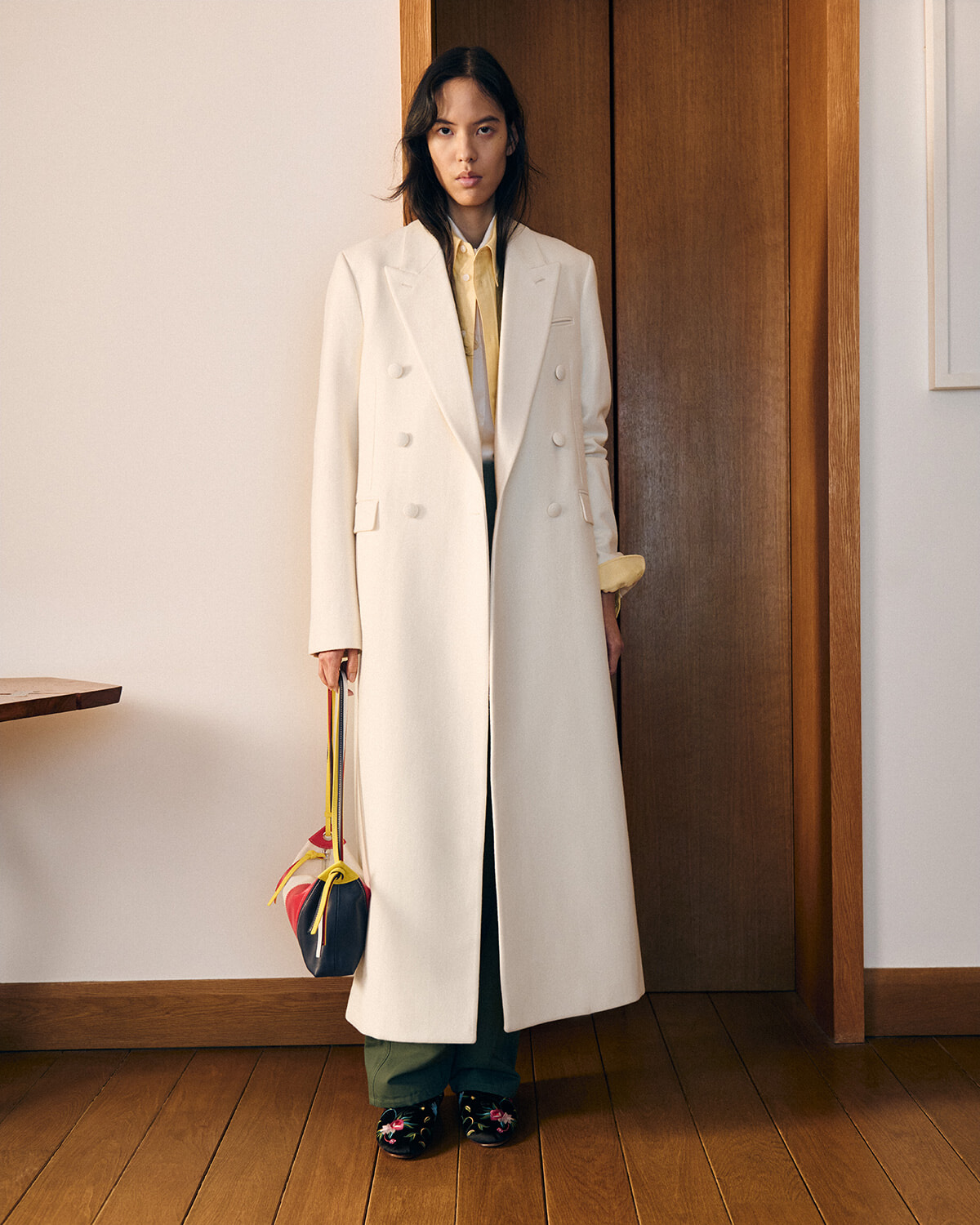 Person standing in a doorway wearing a long cream double-breasted coat, yellow shirt, green pants, and floral shoes, holding a colorful handbag.”,